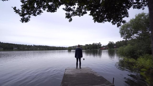 A fashionable young man standing on a pier enjoying the view by a calm lake in Sweden. A steady gimbal footage moving away from the model. A beautiful forest scenery.