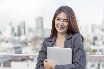 Business Woman Smile and holding computer laptop with office building background.
