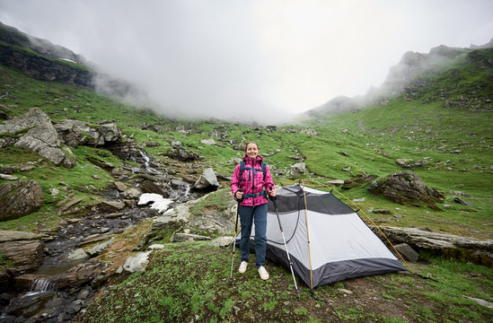 Woman tourist with a backpack and trekking sticks near the tent in the valley of the Romanian mountains. In the background a fog descends on the mountains