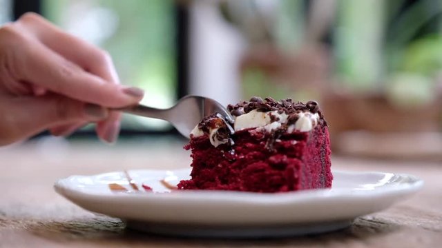 A woman using a fork to cut a piece of red velvet cake with cream and chocolate in white ceramic plate on wooden table