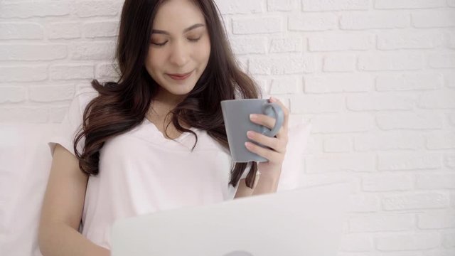 Beautiful Asian woman playing computer or laptop while lying on the bed in her bedroom. Happy female drinking coffee while buying online at home. Lifestyle woman at home concept.