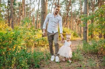 Fototapeta premium Dad and daughter are walking along the forest road in summer sunny park. Stylish man with kid relaxing in forest, having fun. space for text. family vacation