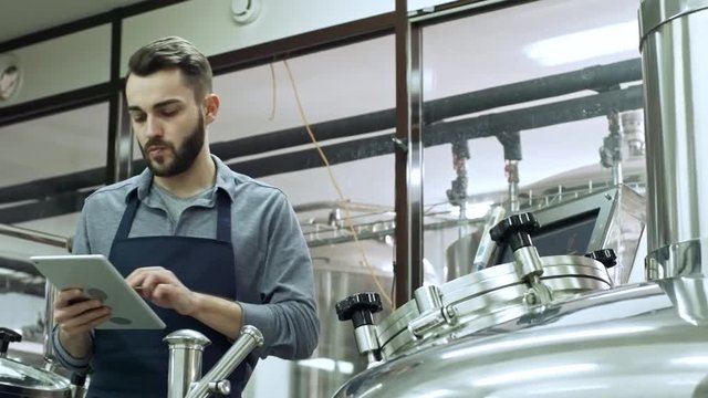Young handsome brewery technician using digital tablet while monitoring the production process at beer plant