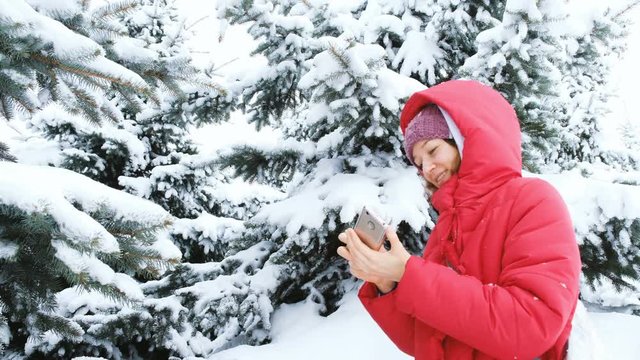 Smiling girl in a down jacket makes selfie on a background of snowy trees in a winter forest