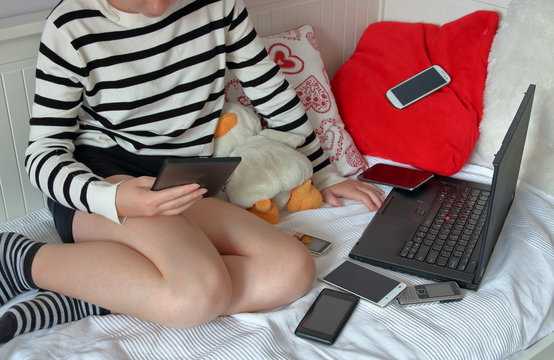 Dziecko (dziewczynka) siedzi na sofie, w ręku trzyma czytnik e-booków, czyta, obok niej leży otwarty laptop i kilka smartfonów, jeden telefon leży na czerwonej poduszce