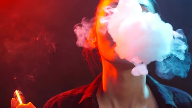 Night life concept. Young woman smokes electronic cigarette in color light