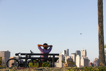 Young man sitting on bench in the park next to his bicycle observing the views of downtown Brisbane city, in Queensland, Australia. Relax after outdoor activity riding bike, success concept