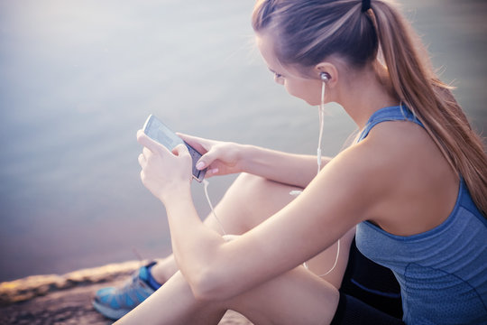 The girl in a t-shirt sitting near the water with a smartphone and headphones after jogging.