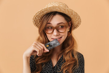 Image of attractive woman 20s wearing straw hat and sunglasses holding and biting credit card,...