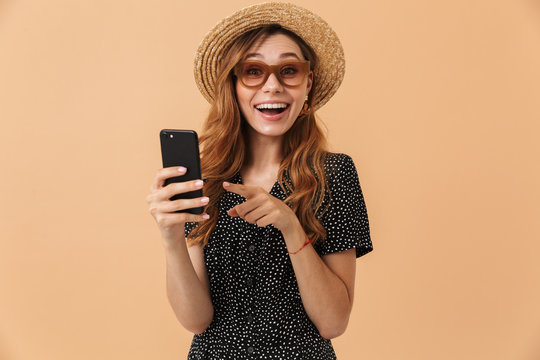 Portrait of surprised joyful woman wearing straw hat and sunglasses laughing and holding smartphone, isolated over beige background