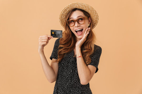 Image of adorable excited woman wearing straw hat and sunglasses rejoicing and holding credit card, isolated over beige background