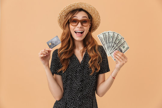 Image of happy rich woman 20s wearing straw hat and sunglasses rejoicing while demonstrating credit card and fan of dollar cash, isolated over beige background