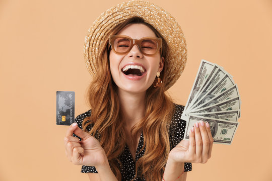 Image of happy young woman 20s wearing straw hat and sunglasses laughing and demonstrating credit card and fan of dollar cash, isolated over beige background