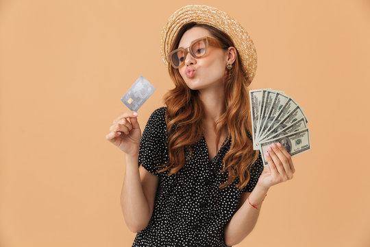 Image of happy rich woman 20s wearing straw hat and sunglasses holding credit card and fan of dollar money, isolated over beige background
