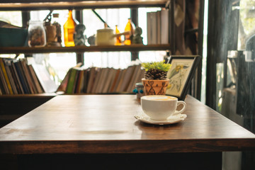 cup of coffee with plants and picture frame place on top of wooden table in living room has bookshelf background