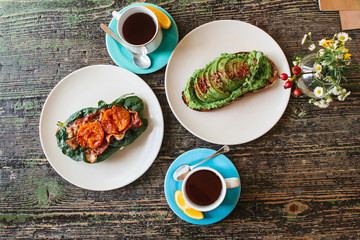 Sandwich with avocado and another sandwich with bacon and tomato next to two cups of tea on a wooden table