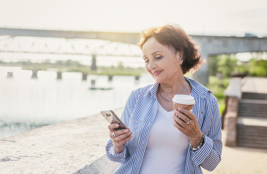 Portrait of a mature attractive stylish woman retired with a cup of coffee and a smartphone in her hands outdoors on a summer sunny day