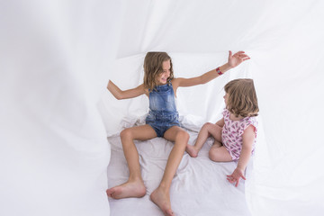 Fototapeta na wymiar Two beautiful sister kids playing under white sheets on bed. Fun indoors. Family love and lifestyle