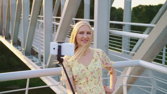 Woman taking selfie on bridge. Cheerful pretty blonde woman standing on pedestrian bridge and taking selfie with monopod in sunset.