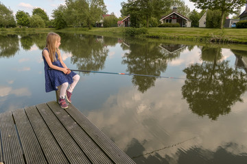 Ein Mädchen sitzt auf einem Pfosten einer Promenade an einem See mit einer Angelrute. Sier wartet, dass ein Fisch anbeißen