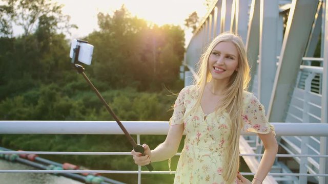 Woman taking selfie on bridge. Cheerful pretty blonde woman standing on pedestrian bridge and taking selfie with monopod in sunset.