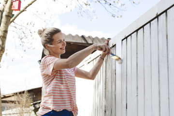 young, pretty woman strokes the garden fence with white paint in summer