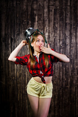 Obraz premium Young funny housewife in checkered shirt and yellow shorts pin up style with kitchen sieve on wooden background.
