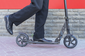 Male legs in trousers and shoes push the scooter along the paving stones near the red wall