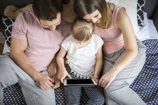 Happy Family Sitting On Bed, Watching Something On Digital Tablet With Little Daughter