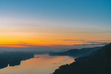 View from high shore on river. Riverbank with forest under thick fog. Gold dawn reflected in water. Yellow glow in picturesque predawn sky. Colorful morning atmospheric landscape of majestic nature.
