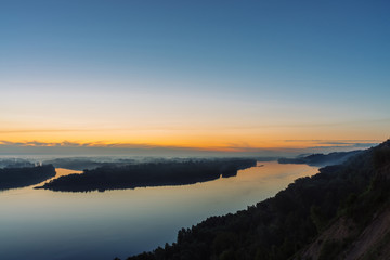 View from high shore on broad river. Riverbank with forest under thick fog. Dawn reflected in water. Yellow glow in picturesque predawn sky. Mystical morning atmospheric landscape of majestic nature.