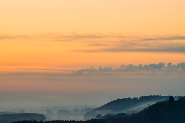 Morning mystic haze above broad valley of river. Gold glow from dawn in sky. Riverbank with forest under fog. Sunlight reflected in water at sunrise. Colorful atmospheric landscape of majestic nature.