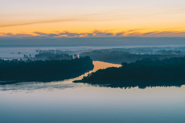 Early blue sky reflected in river water. Riverbank with forest under predawn sky. Yellow stripe in picturesque sky. Fog hid trees on island. Mystical morning atmospheric landscape of majestic nature.