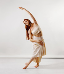 Naklejka premium Dancer posing in studio. Young and beautiful redhead girl in a beige long skirt and top dances and poses in studio. Copy space, gray background