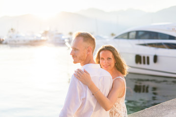 Couple of travelers are sitting in sea resort near yachts during romantic honeymoon vacation at sunset after wedding. Man and woman are happy tourists. Lovers are wearing in white clothes outdoor.