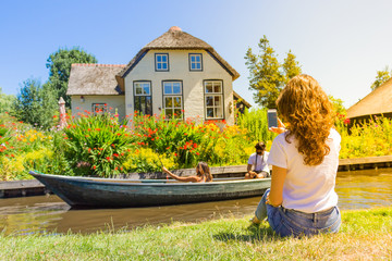 A young woman is photographing a beautiful house in Giethoorn (Netherlands), a couple is floating by