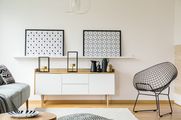 Patterned posters above wooden cupboard in bright living room interior with black armchair. Real photo