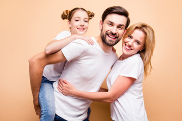 Young adorable attractive beautiful mother of child hugging her husband, bearded handsome man piggy backing their lovely little daughter. Happy family smiling, looking straight on beige background