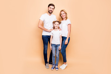 Full length portrait of adorable, attractive family standing together wearing white T-shirts and jeans on beige background. Bearded father and blonde mother keeping their hands on daughter's shoulders