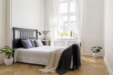 Knit blanket on bed in minimal white bedroom interior with plants and window. Real photo - obrazy, fototapety, plakaty