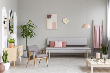 Sofa with pastel pink cushions in real photo of grey living room interior with retro armchair, fresh green plants, simple poster and blanket hanging on ladder