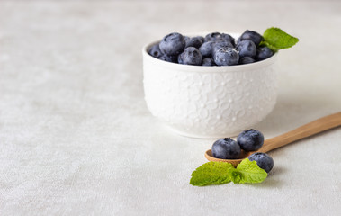 Fresh blueberries in a white vintage ceramic bowl and wooden spoon with mint on light grey background. Copy space