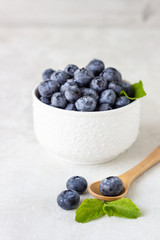 Fresh blueberries in a white vintage ceramic bowl and wooden spoon with mint on light grey background. Copy space