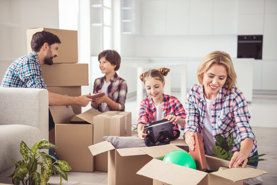 Young happy smiling family four persons unpacking carton boxes with stuff in light kitchen living room, moving to new flat