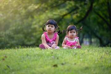 Naklejka na ściany i meble Little sister play and sit on the green lawn in park. Cute girls relax in park.
