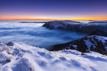 Amazing view from top of mountain down to valley filled with low clouds, inversion. Colorful sunset light. Snow covered winter landscape. Adventure, hike, travel, trek, explore, discover.