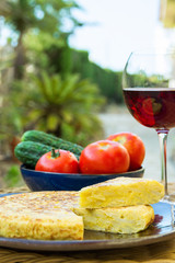 Traditional Spanish Omelet Tortilla with Potatoes Eggs Cut out Wedge Organic Vegetables Tomatoes Cucumbers Glass of Red Wine on Rattan Table in Garden Outdoors. Summer Day. Lifestyle