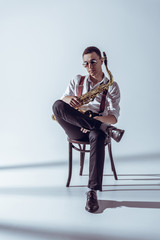stylish young performer in sunglasses sitting on chair and holding saxophone on grey