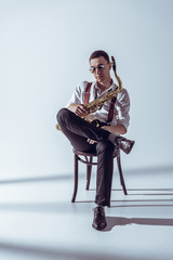 Fototapeta premium stylish young performer in sunglasses sitting on chair and holding saxophone on grey