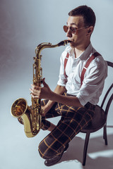 Fototapeta na wymiar high angle view of stylish young performer sitting on chair and playing saxophone on grey